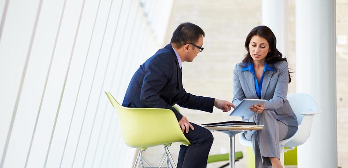 A man and woman discuss business collaborating on an online project using a touchpad tablet in a modern office space. (Photo: AdobeStock)