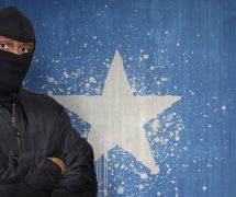 A man posing as an Islamic terrorist in a mask standing in front of a wall painted with the national flag of Somalia. (Photo: AdobeStock)