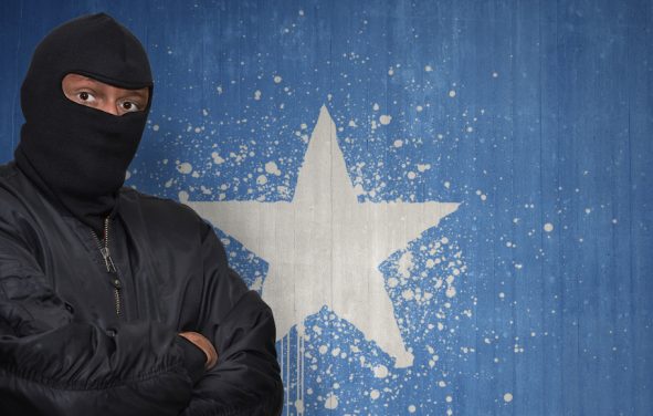 A man posing as an Islamic terrorist in a mask standing in front of a wall painted with the national flag of Somalia. (Photo: AdobeStock)