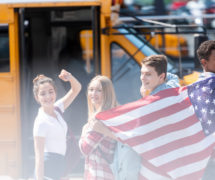 A group of American public school teenage students with an American flag in front of school bus. (Photo: AdobeStock)