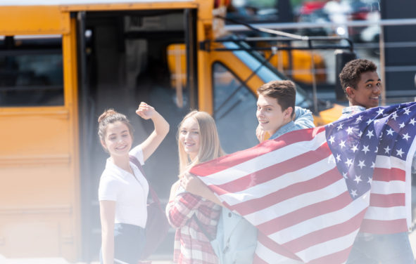 A group of American public school teenage students with an American flag in front of school bus. (Photo: AdobeStock)