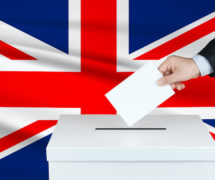 Graphic concept of an election in Great Britain, depicting by the hand of a voter putting his vote in the ballot box with the flag of the United Kingdom in the background. (Photo: AdobeStock)