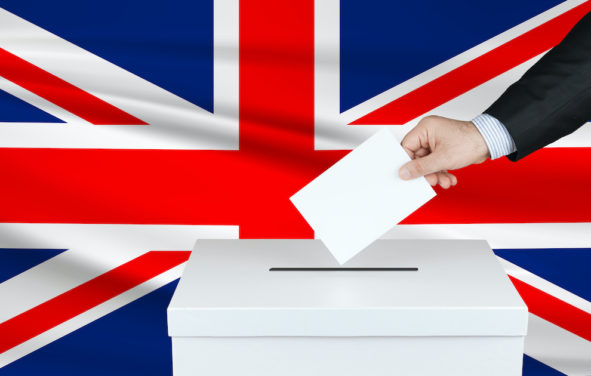 Graphic concept of an election in Great Britain, depicting by the hand of a voter putting his vote in the ballot box with the flag of the United Kingdom in the background. (Photo: AdobeStock)