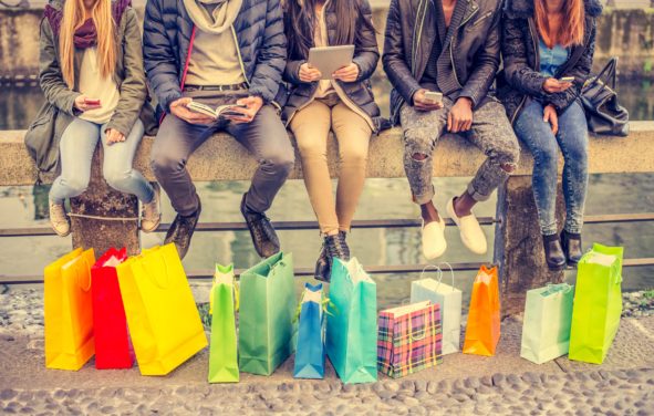 Group of friends sitting outdoors with shopping bags; several people holding smartphones and tablets. (Photo: AdobeStock/ OneInchPunch/PPD)