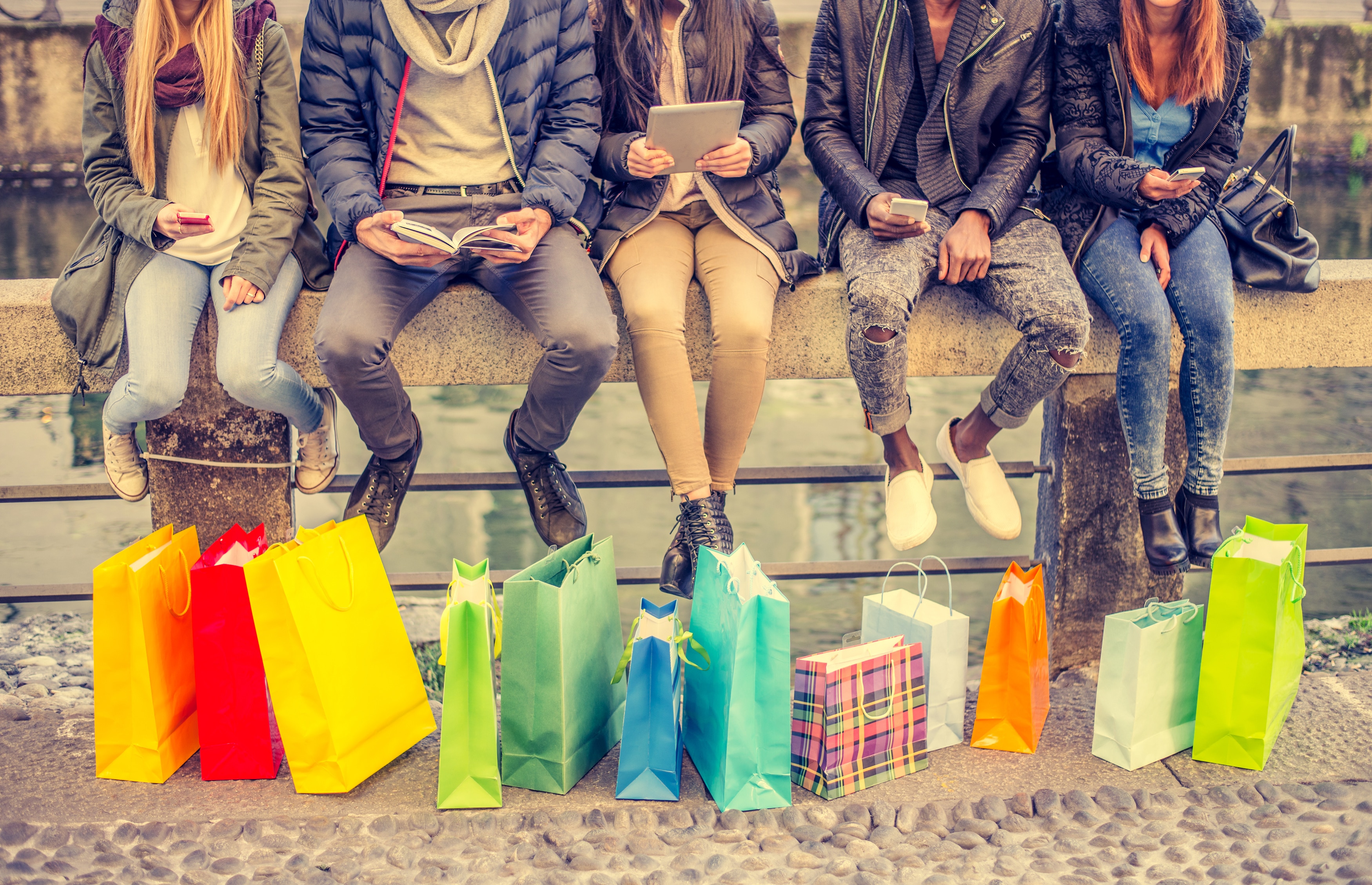 Group of friends sitting outdoors with shopping bags; several people holding smartphones and tablets. (Photo: AdobeStock/ OneInchPunch/PPD)